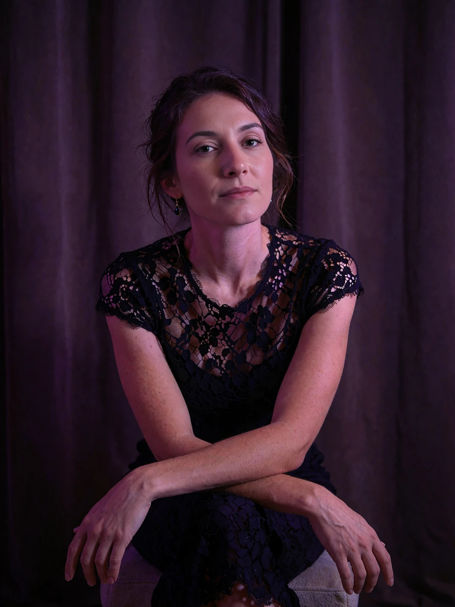 Woman with dark hair pinned back in a black lace evening top seated on a low stool in soft light