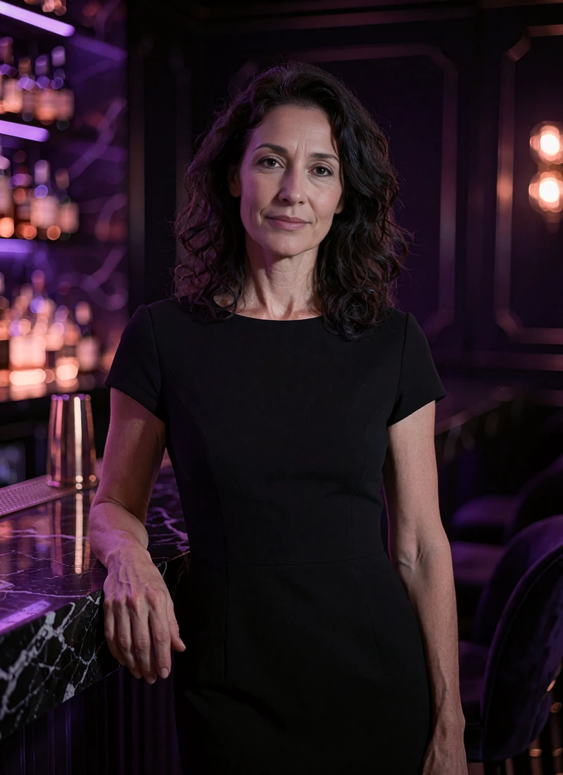 Elegant woman in her mid forties with wavy dark hair standing beside a marble bar counter with amber light behind her