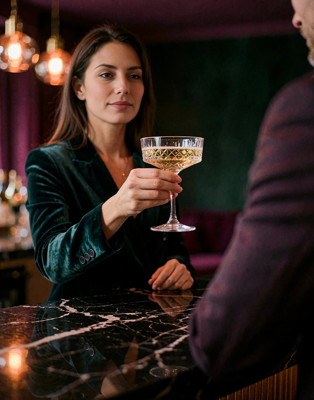 Poised woman in a dark emerald velvet blazer raising a crystal champagne coupe at a black marble cocktail bar